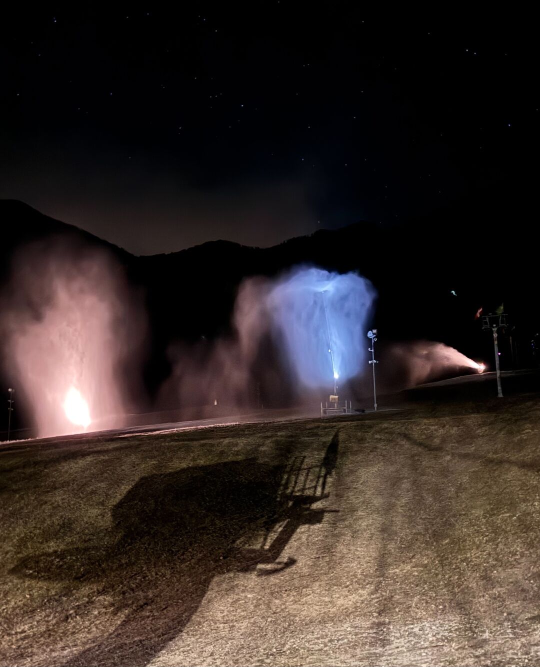 Paesaggio notturno, con cannoni che sparano neve su una pista, in lontananza catena di monti con cielo stellato